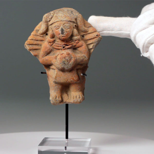 Pre-Columbian ceramic figure adorned with an ornate collar, ear pieces, and headress with depicted horns. The figure is holding a ball of some sort at its waist over clothing. Behind the human figure is a large horizontally ridged structure that looks as if it was larger at some point but has been broken off making it into a more diamond shape.