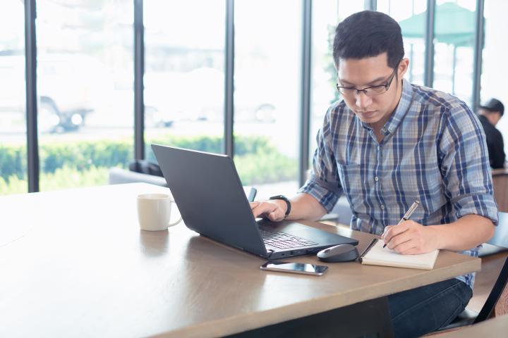 man typing on laptop and writing something down, with a coffee cup beside him